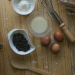 A flat lay of baking essentials including eggs, flour, chocolate chips, and a whisk on a wooden surface.