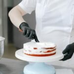 A chef in gloves and mask carefully decorates a cake with white cream frosting in a kitchen.